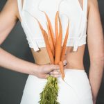 A person in white holding a bunch of fresh carrots against a gray background, symbolizing health and wellness.