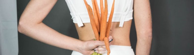 A person in white holding a bunch of fresh carrots against a gray background, symbolizing health and wellness.