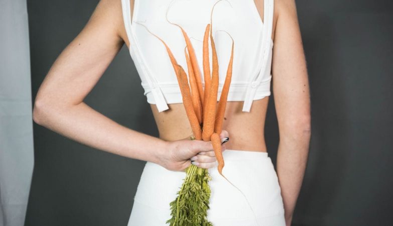 A person in white holding a bunch of fresh carrots against a gray background, symbolizing health and wellness.