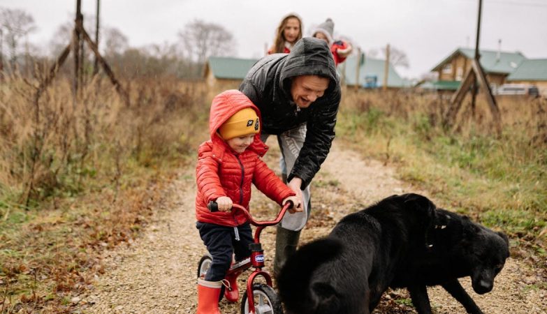 Dad helps son ride a bike on a path with family and dog, capturing a joyful outdoor moment.